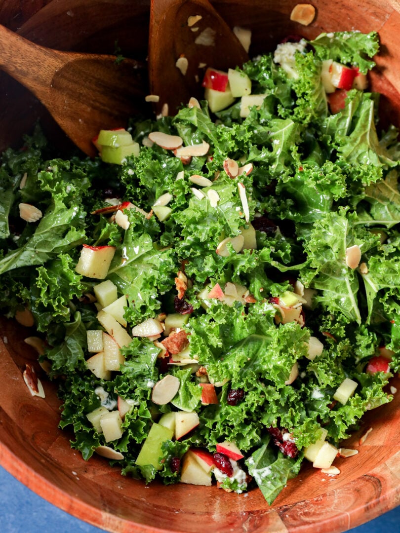 Apple and kale salad with chopped apples, dried cranberries, and sliced almonds in a wooden bowl with serving spoons.