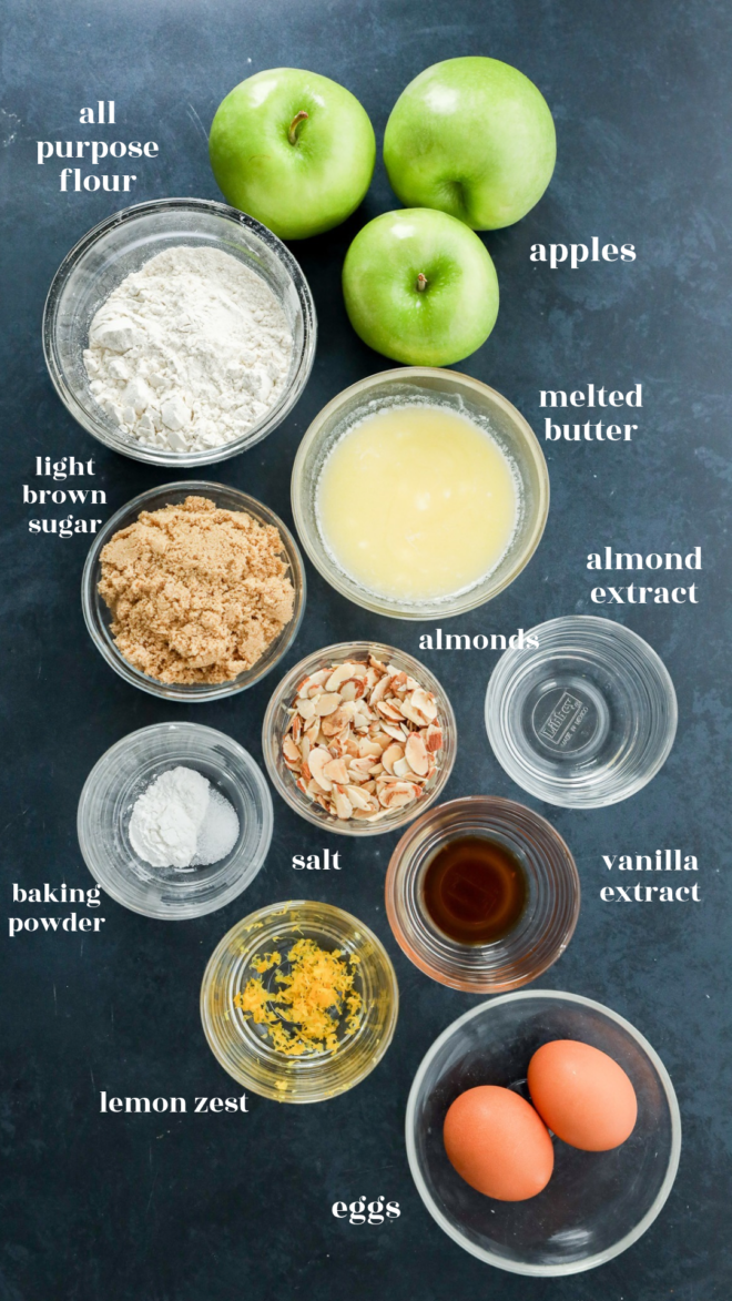 A top-down view of ingredients for a recipe, including flour, apples, butter, brown sugar, sliced almonds, baking powder, salt, vanilla extract, lemon zest, and eggs in individual bowls.
