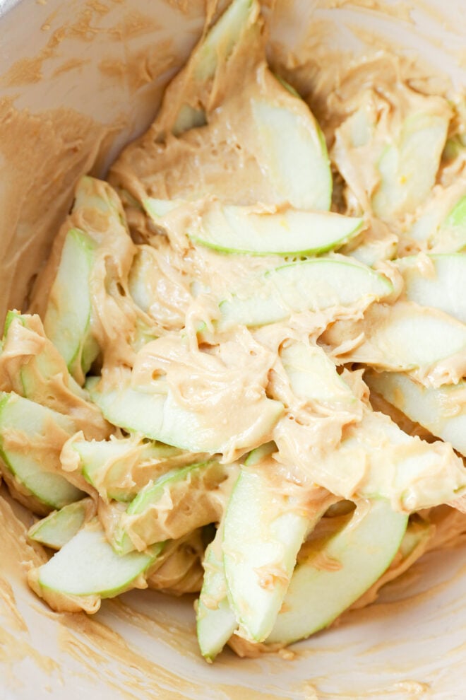 Thin slices of green apple coated in creamy peanut butter in a mixing bowl.