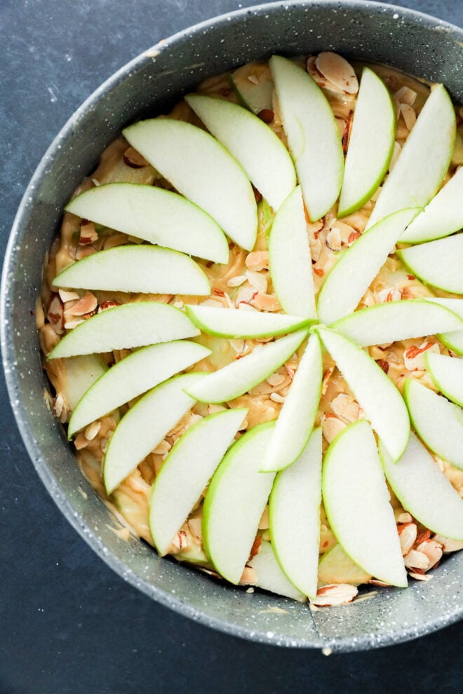 A round cake pan containing unbaked batter topped with a decorative pattern of sliced green apples and almond slivers.
