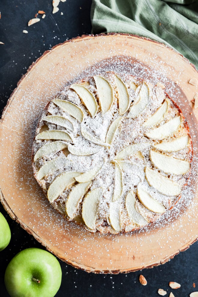 Apple cake topped with sliced apples and powdered sugar on a round wooden serving board.