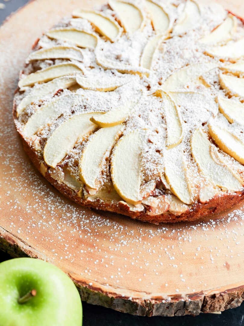 An apple cake topped with sliced apples and powdered sugar, served on a wooden platter next to a green apple.