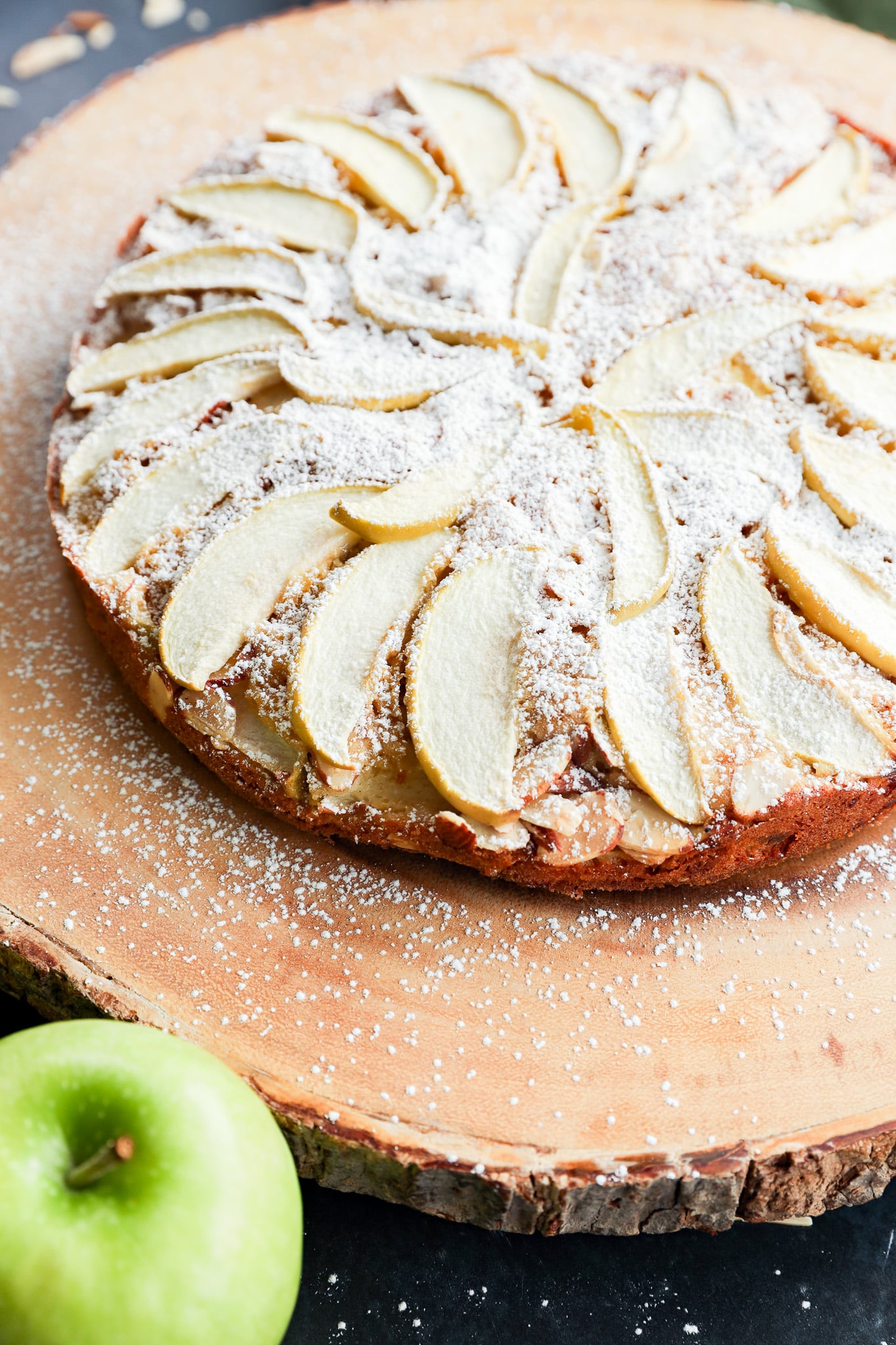 An apple cake topped with sliced apples and powdered sugar, served on a wooden platter next to a green apple.