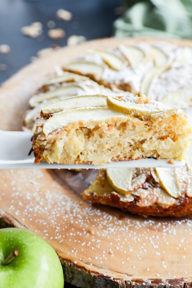 A slice of apple cake topped with sliced apples and powdered sugar being lifted from a larger cake on a wooden board.