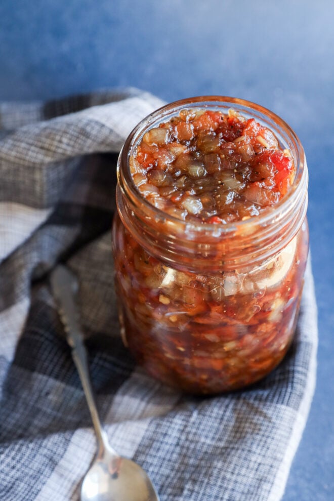 A glass jar filled with chunky bacon onion jam, resting on a plaid cloth next to a silver spoon.