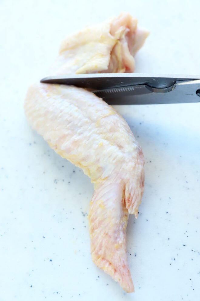 Kitchen shears cutting a raw chicken wing on a cutting board.