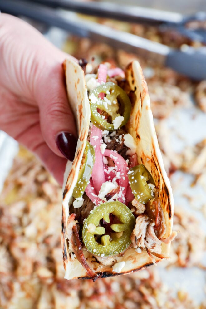 A hand holds an instant pot carnitas taco on a flour tortilla, topped with shredded meat, pickled onions, jalape&ntilde;o slices, and crumbled cheese.