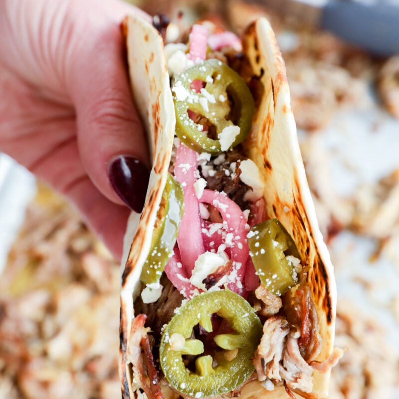 A hand holds an instant pot carnitas taco on a flour tortilla, topped with shredded meat, pickled onions, jalapeño slices, and crumbled cheese.