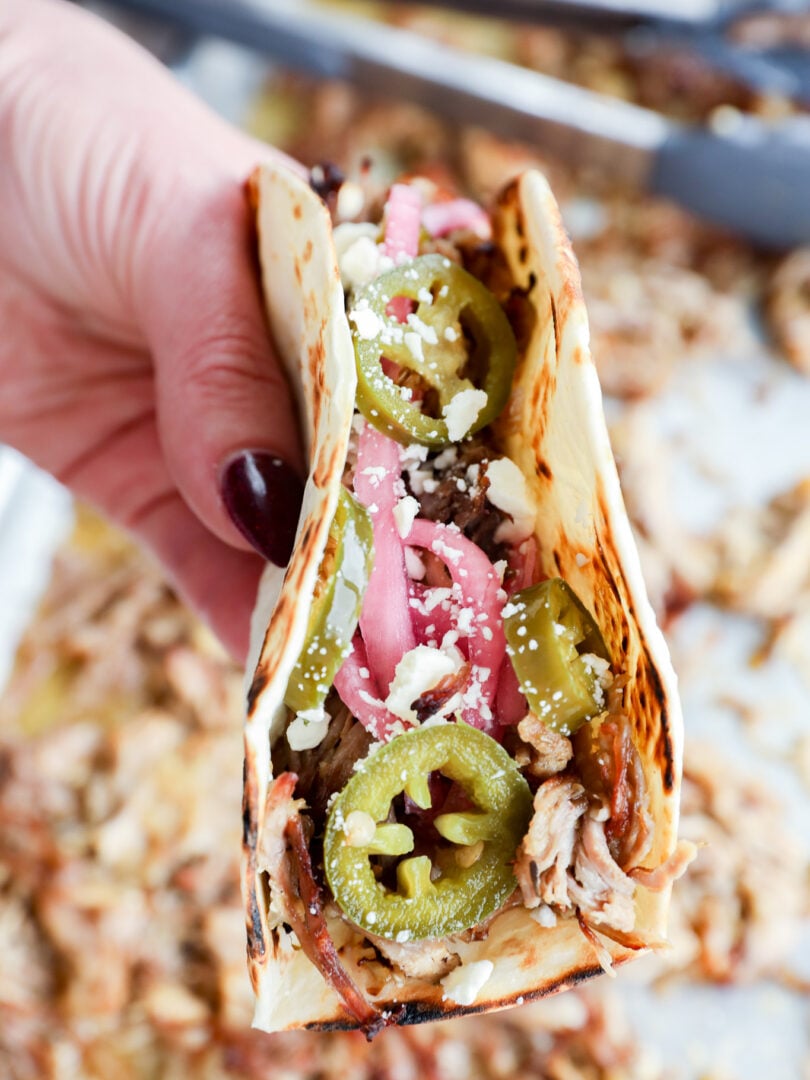 A hand holds an instant pot carnitas taco on a flour tortilla, topped with shredded meat, pickled onions, jalapeño slices, and crumbled cheese.