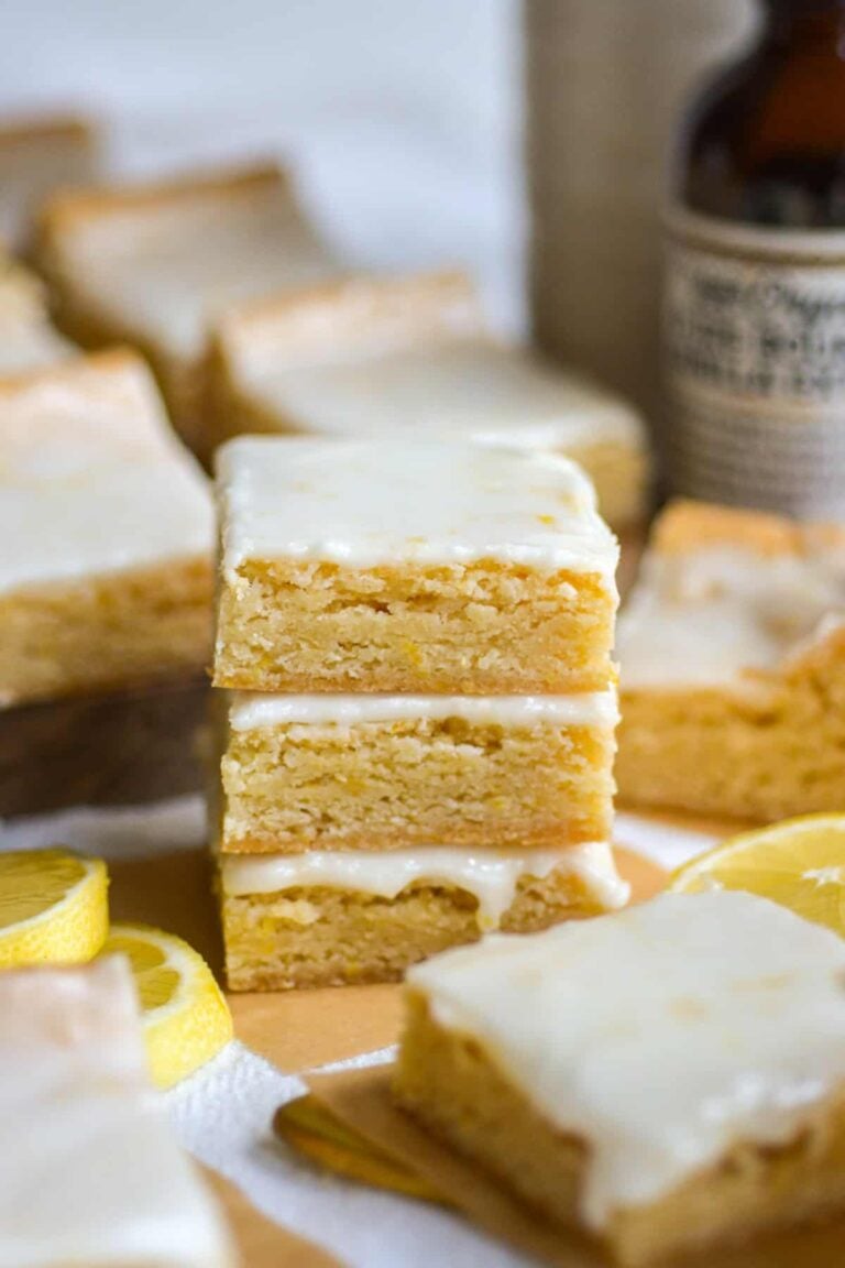 A stack of three lemon bars with white glaze, surrounded by more lemon bars and sliced lemons.