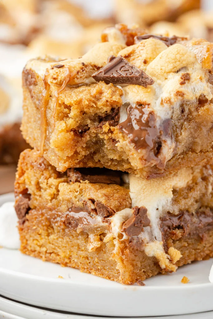 Two stacked smores cookie bars with melted chocolate, toasted marshmallows, and caramel drizzle on a white plate.