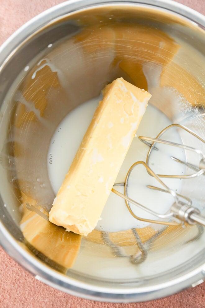 A stick of butter, a splash of milk, and a metal whisk attachment inside a mixing bowl.