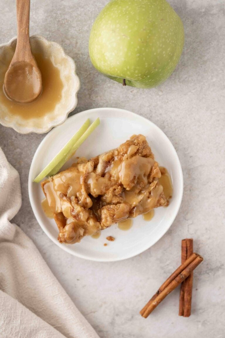 A slice of apple cake topped with caramel sauce on a white plate, next to a whole green apple, a bowl of caramel, and two cinnamon sticks.